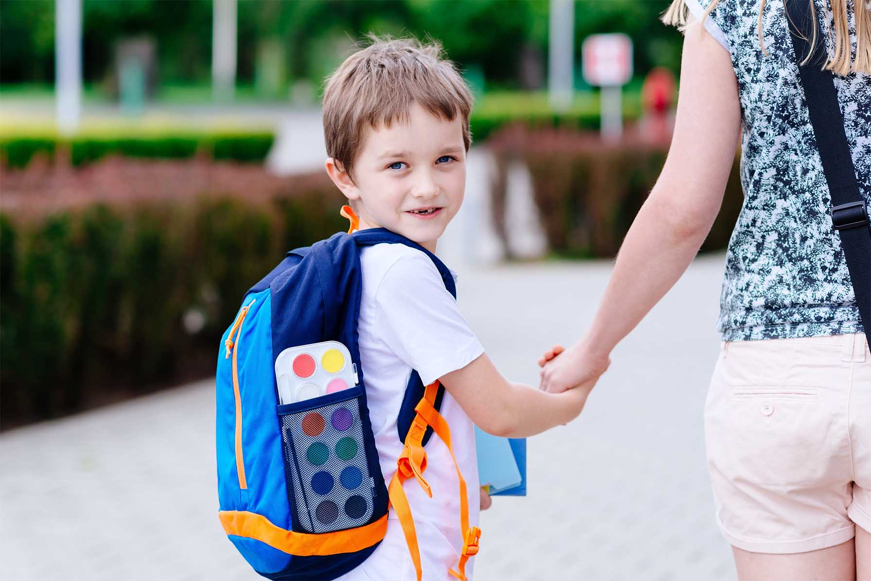 A little boy wearing a back-pack is holding his mom's hand as he turns and smiles at the camera.